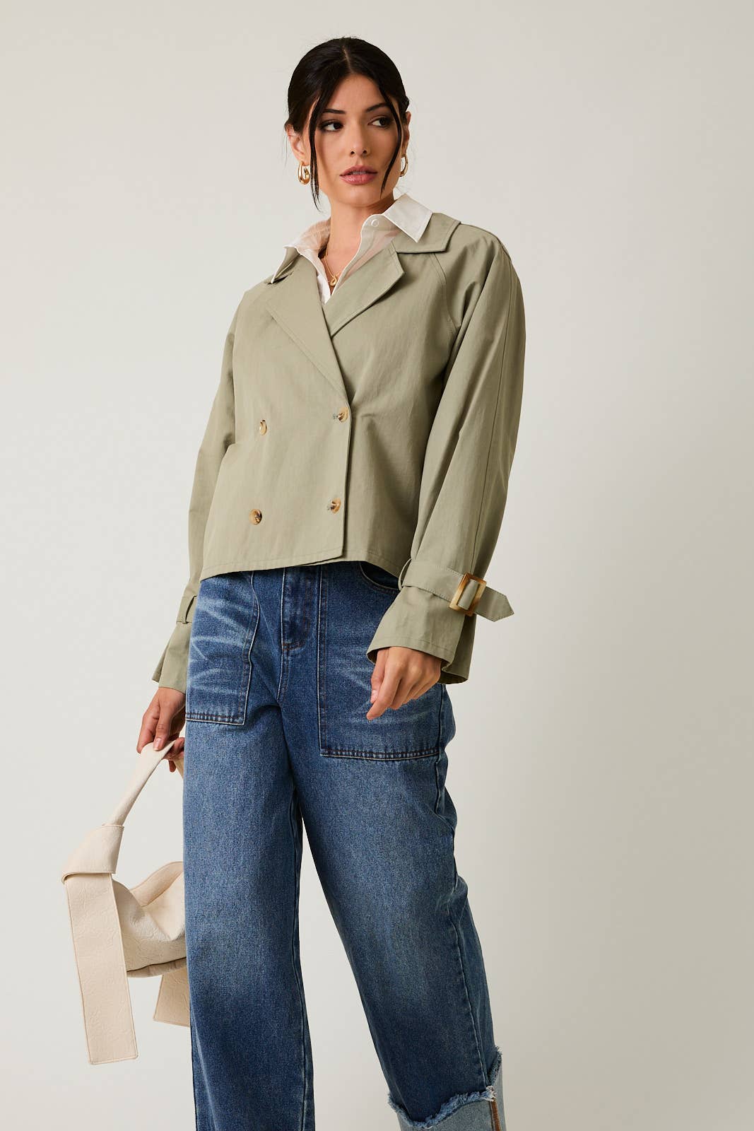 Woman wearing a sage green cropped trench jacket and blue jeans holding a beige handbag on a plain background