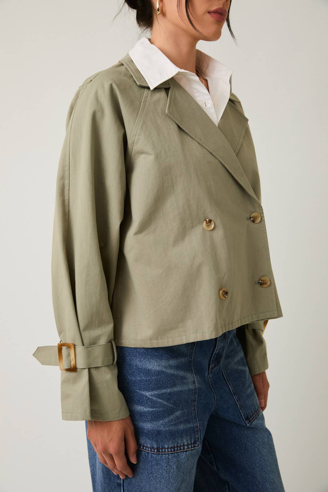 Woman wearing a sage green cropped trench jacket with tortoise shell buttons on a white background