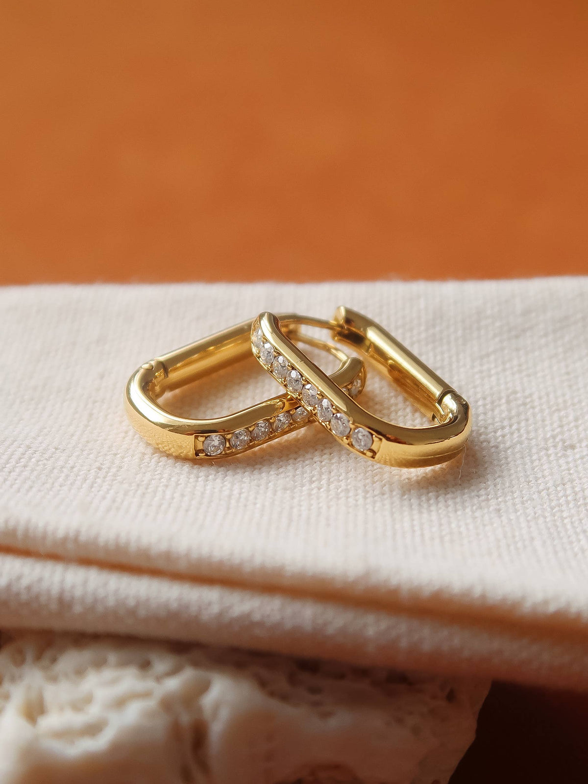 Two gold huggie hoop earrings with pavé-set clear stones, resting on a folded cream fabric surface against a warm orange-brown background.