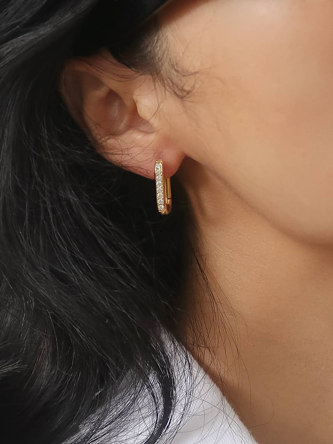 A close-up of a model's ear wearing a small gold huggie hoop earring adorned with clear pavé stones.