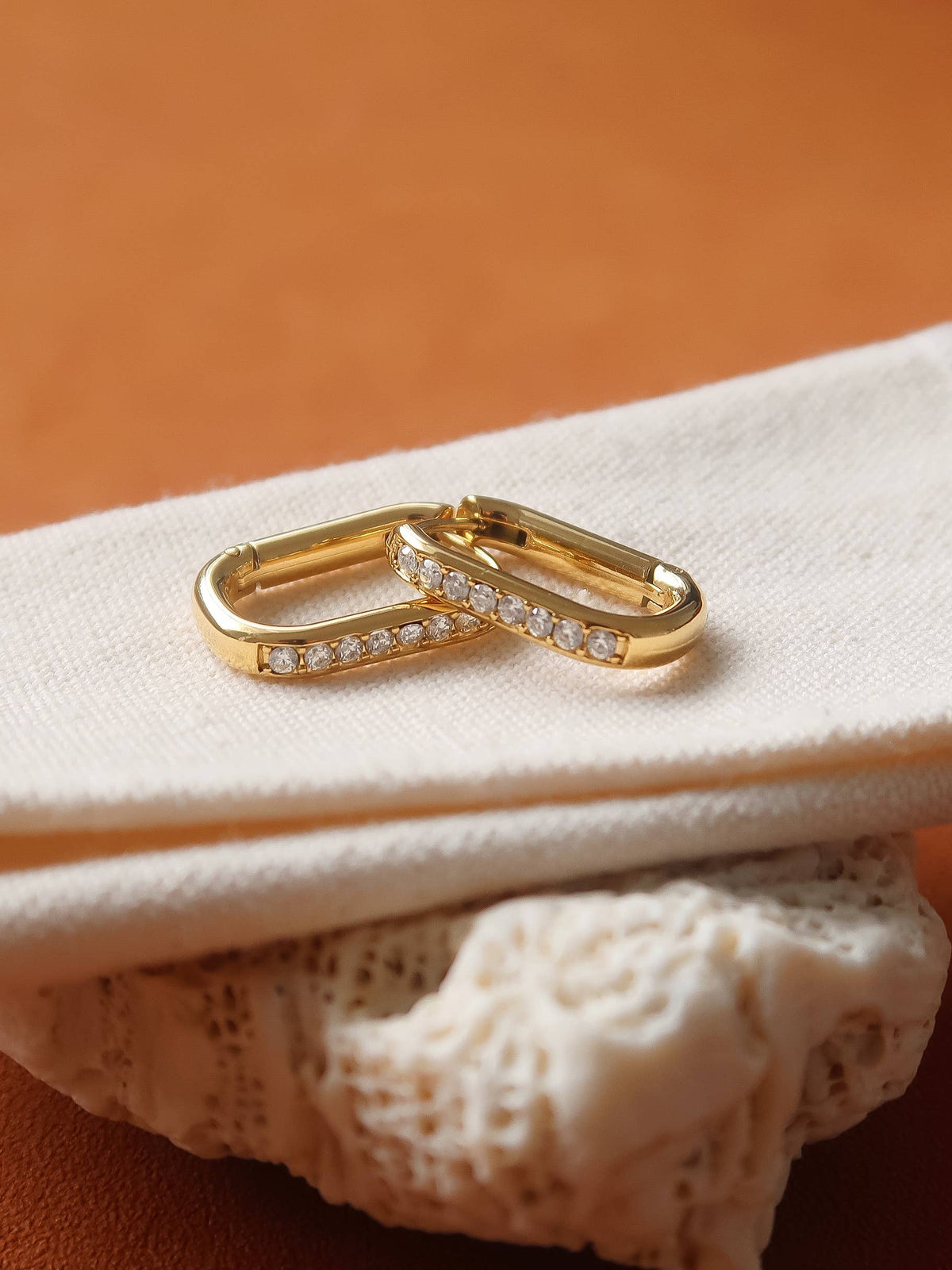 A pair of gold huggie hoop earrings with a row of small clear stones, resting on a folded cream fabric surface with a textured beige object below and a warm brown background.