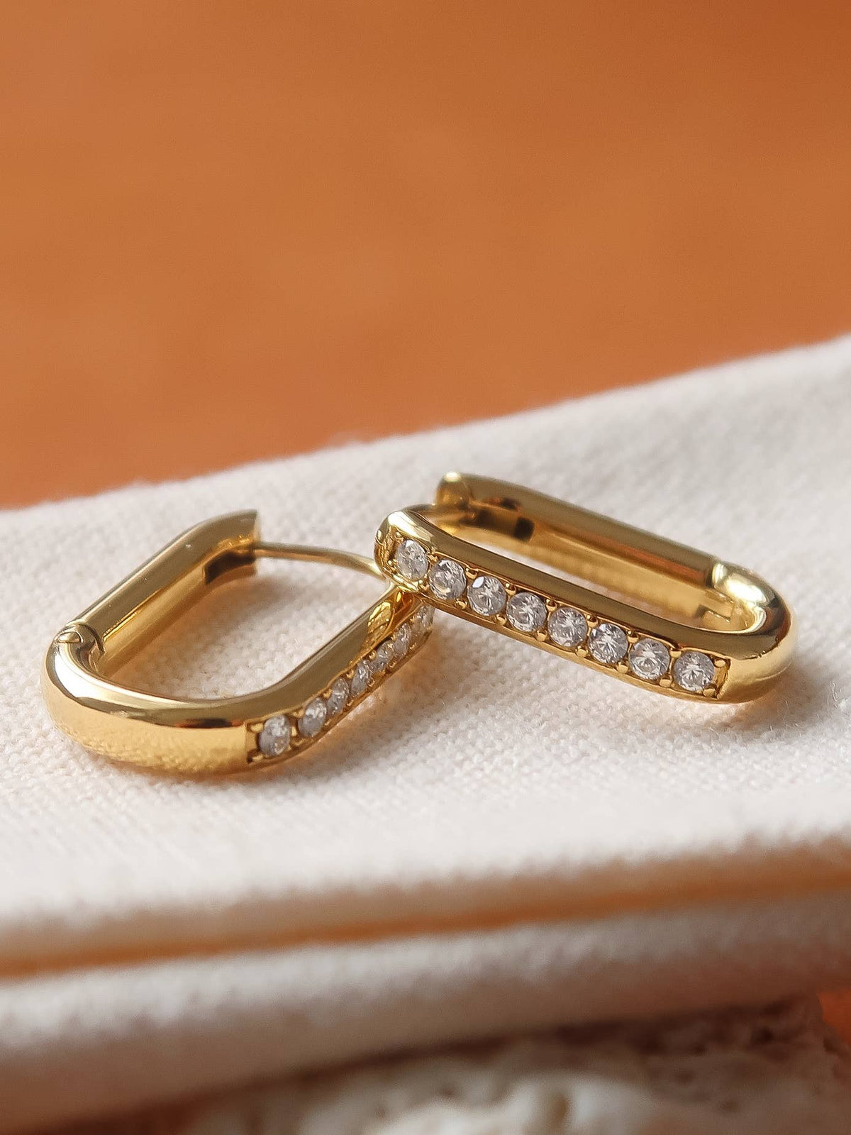 A pair of gold hoop earrings with small clear stones set along the front, resting on a textured cream fabric against a warm orange-brown background.
