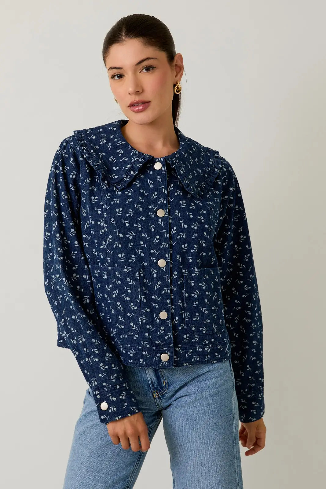 Woman wearing a medium denim blue floral patterned jacket and light blue jeans on a plain background