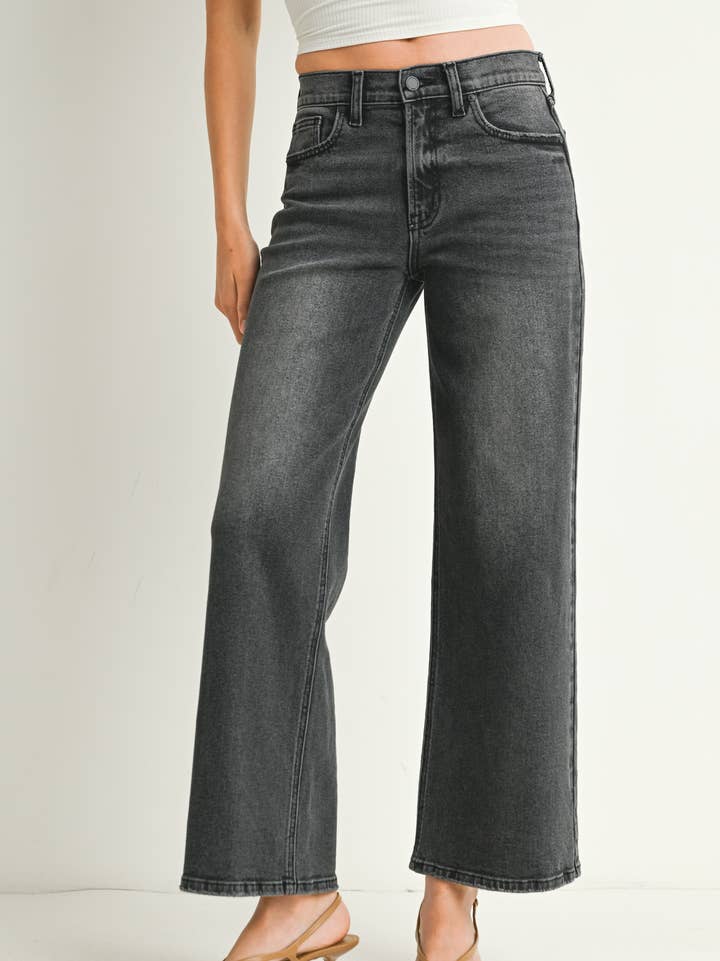 A woman wearing a pair of washed grey straight leg jeans, paired with a white crop top and beige flat sandals, standing against a plain off-white studio background.