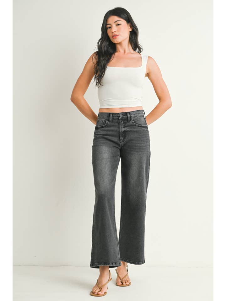 A woman wearing a pair of washed grey straight leg jeans, paired with a white crop top and beige flat sandals, standing against a plain off-white studio background.