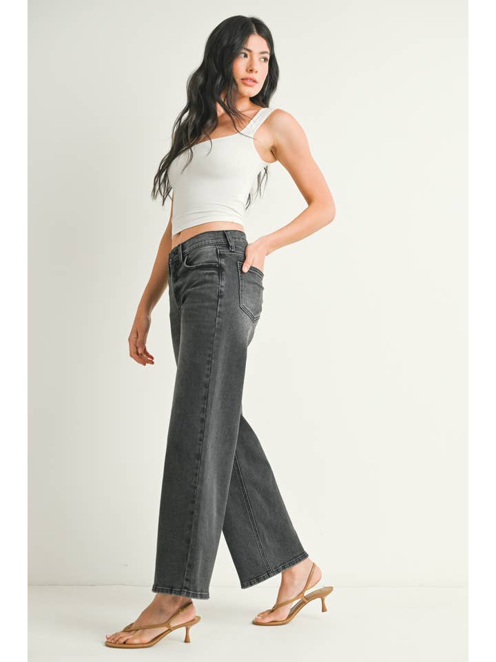 A woman wearing a pair of washed grey straight leg jeans, paired with a white crop top and beige flat sandals, standing against a plain off-white studio background.