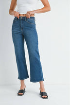 A woman wearing a pair of mid rise dark denim slim wide leg jeans, styled with a white ribbed crop top and black slide sandals, set against a plain white studio background.