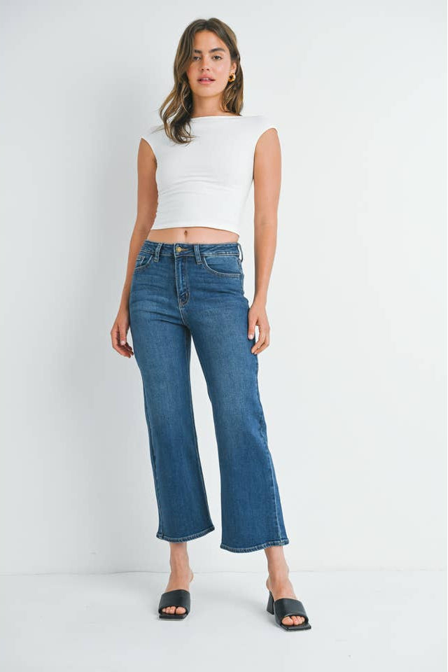 A woman wearing a pair of mid rise dark denim slim wide leg jeans, styled with a white ribbed crop top and black slide sandals, set against a plain white studio background.