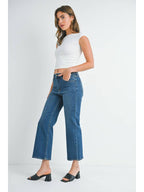 A woman wearing a pair of mid rise dark denim slim wide leg jeans, styled with a white ribbed crop top and black slide sandals, set against a plain white studio background.