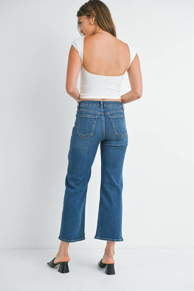 Back view of a woman wearing a pair of mid rise dark denim slim wide leg jeans, styled with a white ribbed crop top and black slide sandals, set against a plain white studio background.