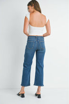 Back view of a woman wearing a pair of mid rise dark denim slim wide leg jeans, styled with a white ribbed crop top and black slide sandals, set against a plain white studio background.