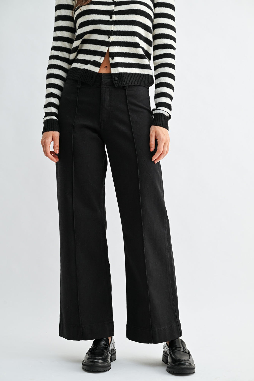 A woman wearing black wide-leg trouser jean, a black and white striped cardigan, and black leather loafers, photographed against a light gray studio background.