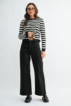 A woman wearing black wide-leg trouser jean, a black and white striped cardigan, and black leather loafers, photographed against a light gray studio background.
