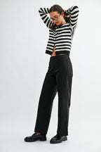 A woman wearing black wide-leg trouser jean, a black and white striped cardigan, and black leather loafers, photographed against a light gray studio background.