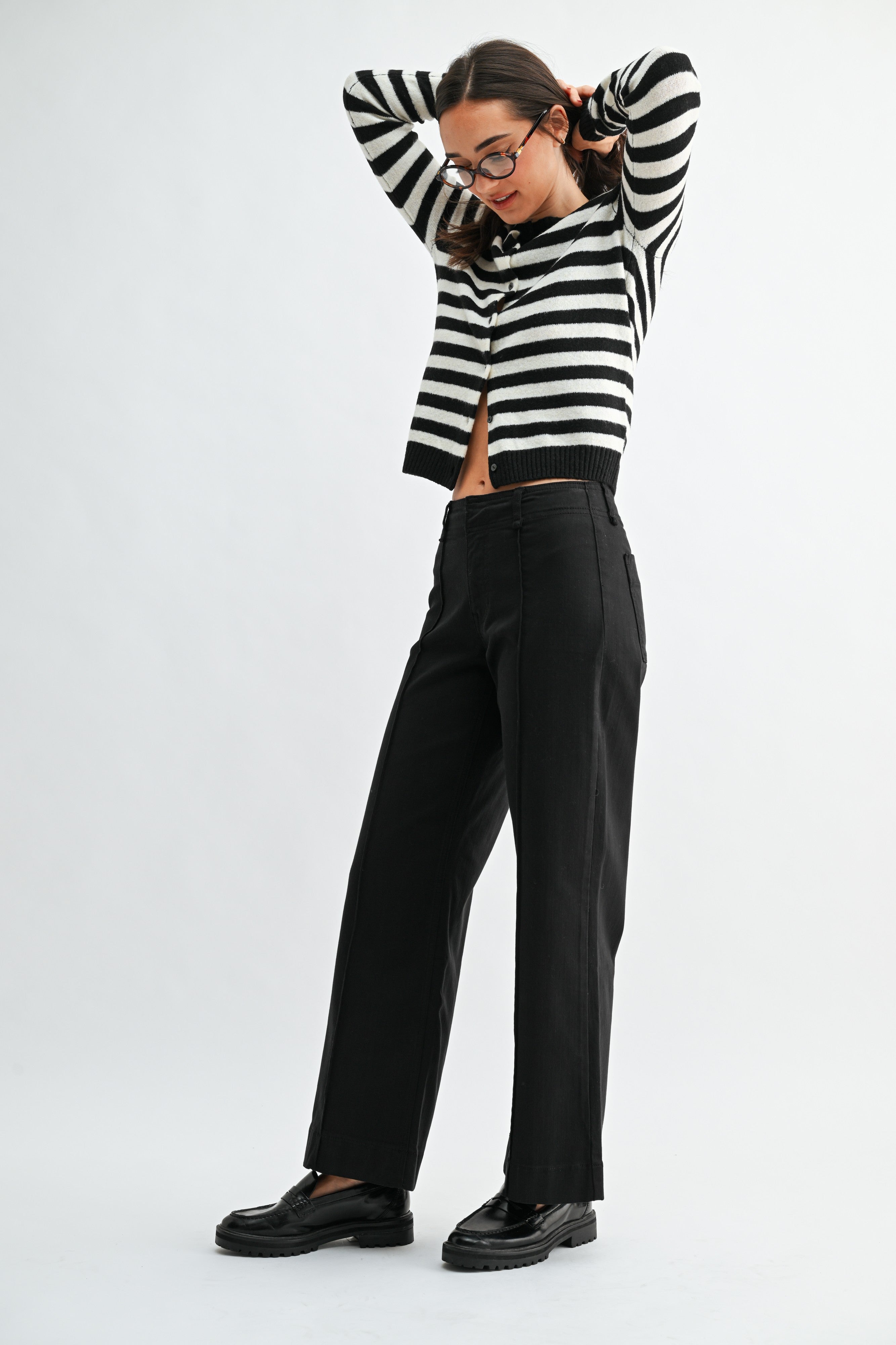 A woman wearing black wide-leg trouser jean, a black and white striped cardigan, and black leather loafers, photographed against a light gray studio background.