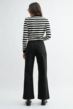 Back view of a woman wearing black wide-leg trouser jean, a black and white striped cardigan, and black leather loafers, photographed against a light gray studio background.
