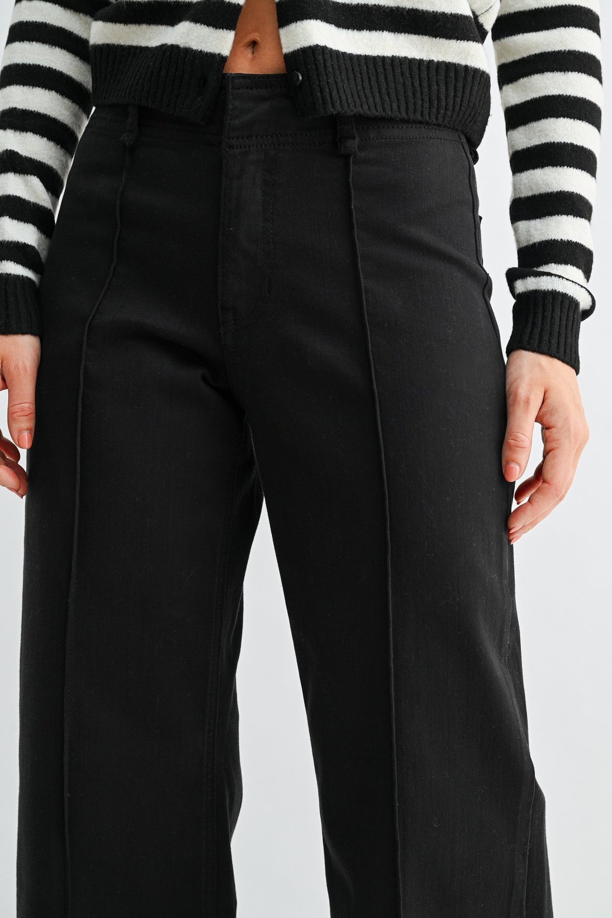 A close-up of a woman wearing black wide-leg twill trouser jeans and a black-and-white striped cropped sweater, cropped from the chest down against a light gray studio background.