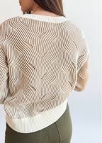 Back view of a woman wearing a mocha and cream two-tone sweater design standing against a brick wall.