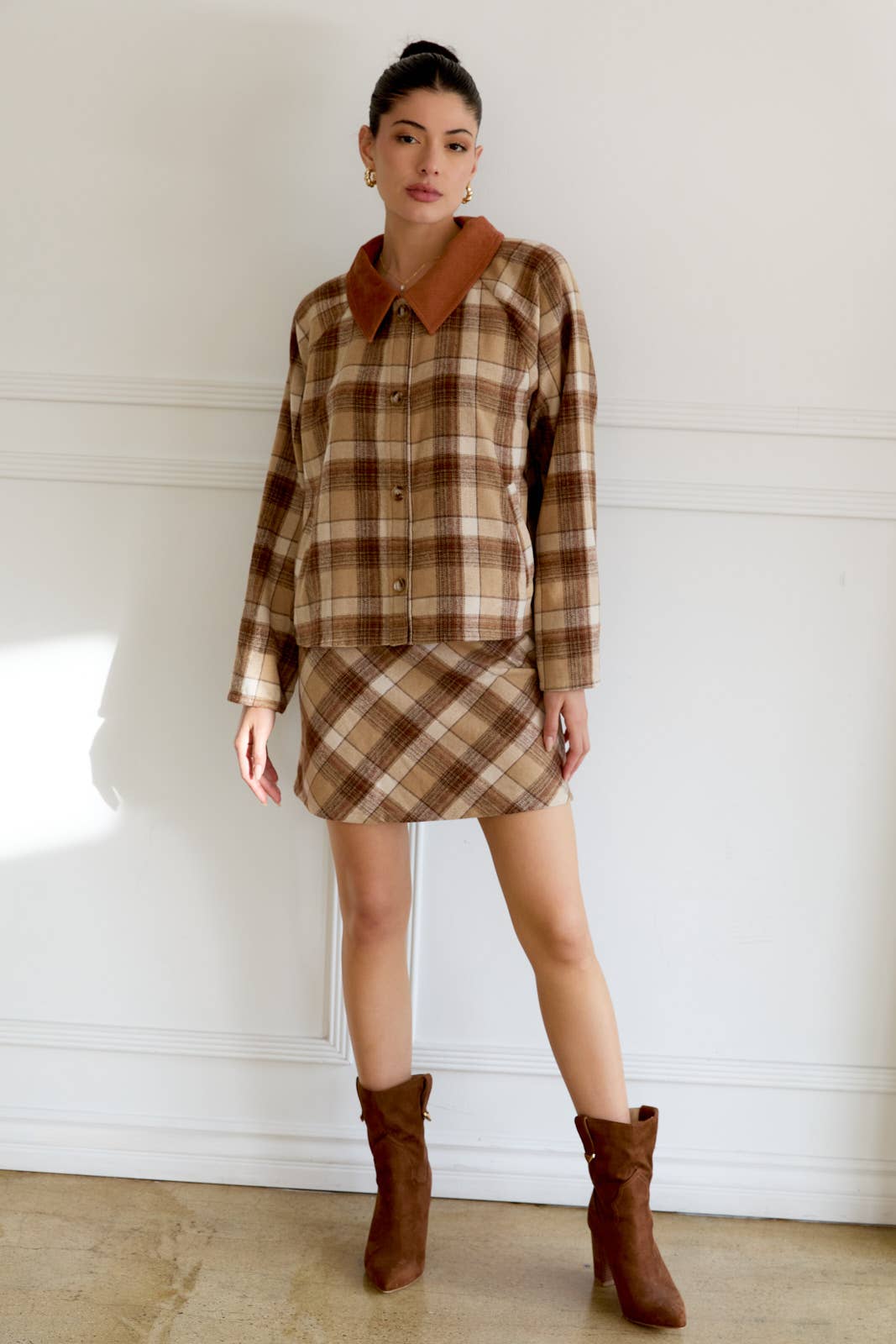 Woman wearing a plaid top with a brown skirt, standing against a white wall.