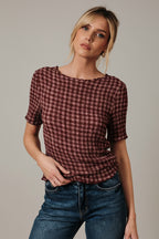 A woman wearing a short-sleeve burgundy and mauve gingham popcorn texture stretch top with blue jeans, posing in front of a draped white fabric backdrop.
