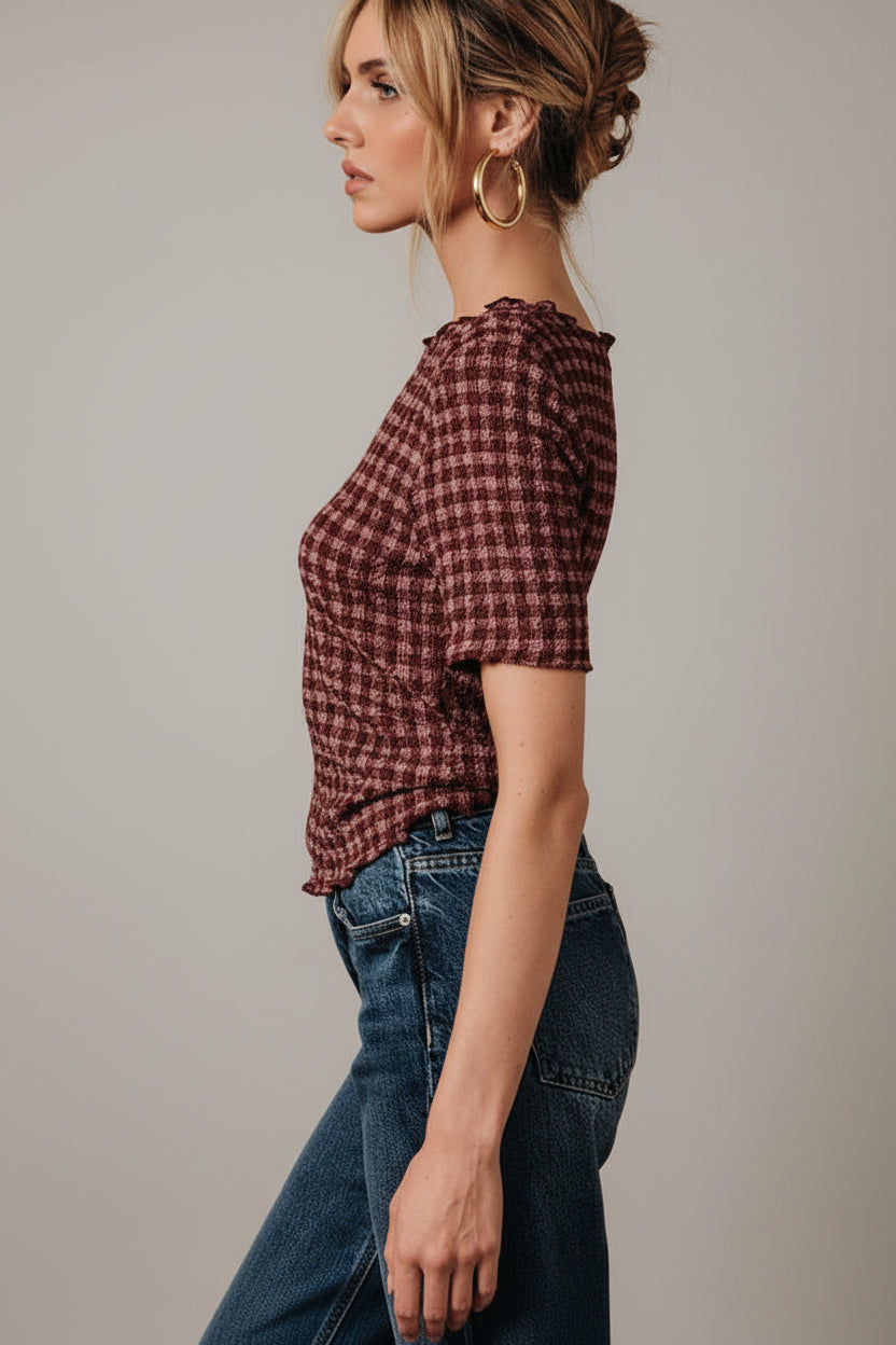 A woman wearing a short-sleeve burgundy and mauve gingham popcorn texture stretch top with blue jeans, posing in front of a draped white fabric backdrop.