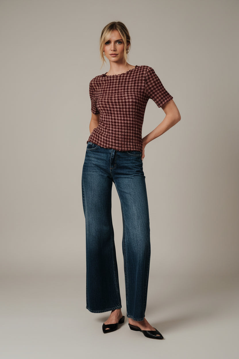 A woman wearing a short-sleeve burgundy and mauve gingham popcorn texture stretch top with blue jeans, posing in front of a draped white fabric backdrop.