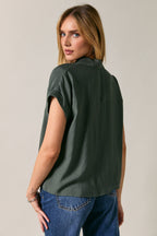 A woman wearing a dark green short-sleeve button-up shirt, shown from the back, paired with blue jeans against a plain light gray studio background.