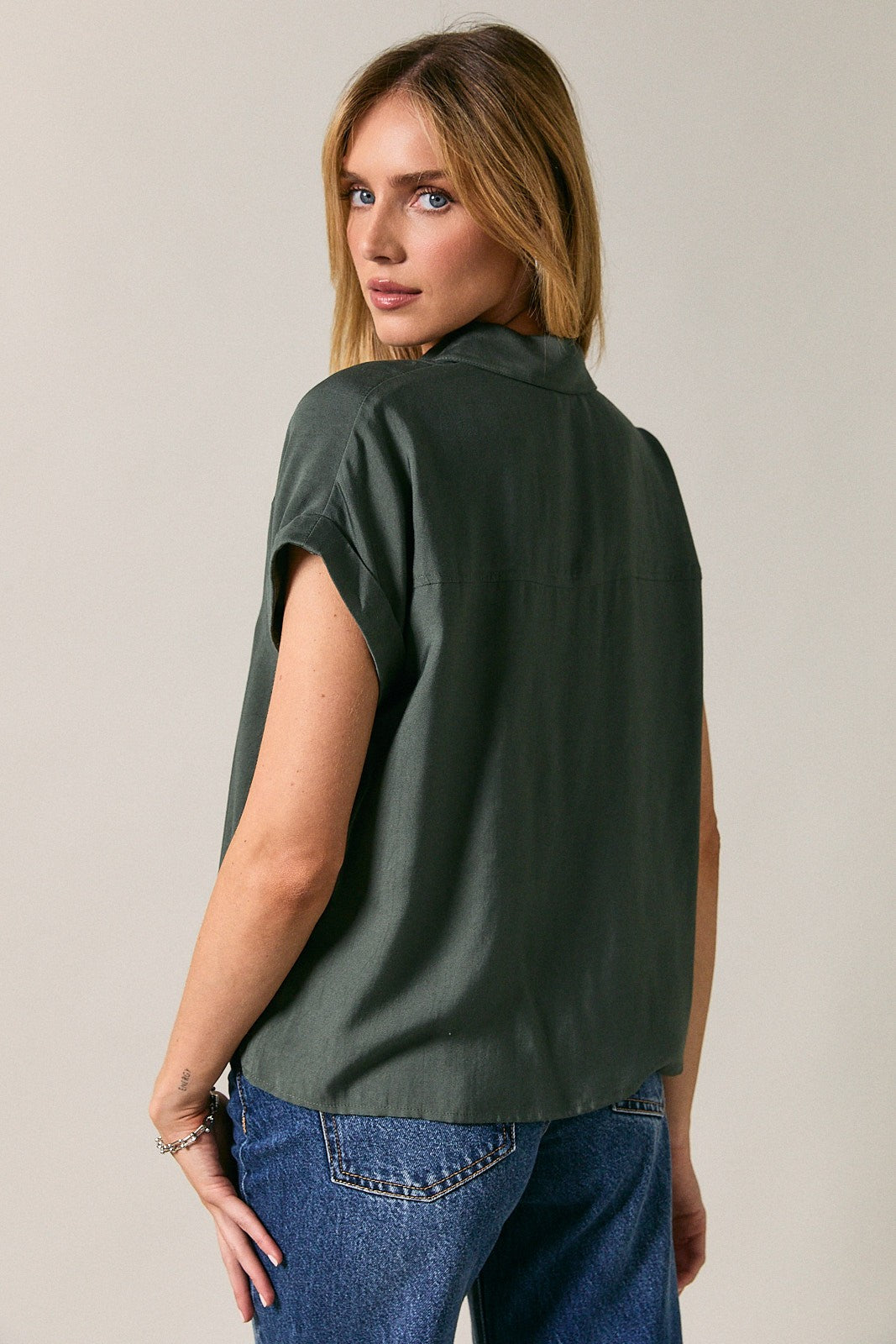 A woman wearing a dark green short-sleeve button-up shirt, shown from the back, paired with blue jeans against a plain light gray studio background.