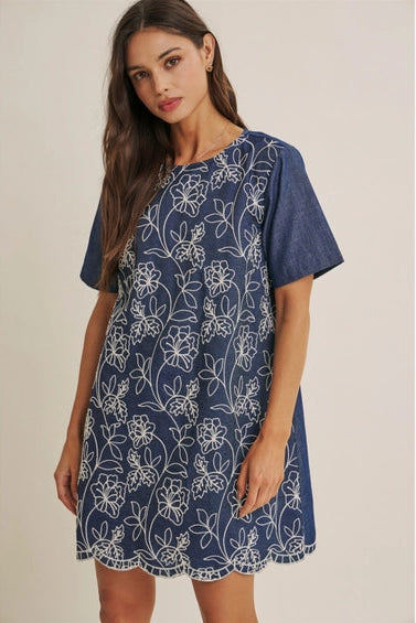 A woman wearing a short-sleeve blue dress with a white floral embroidered pattern, set against a plain beige studio background.