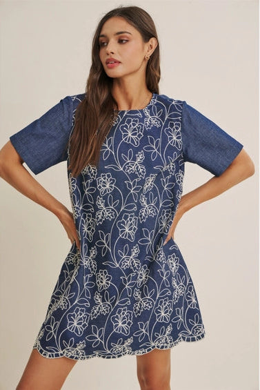 A woman wearing a short-sleeve blue dress with a white floral embroidered pattern, set against a plain beige studio background.
