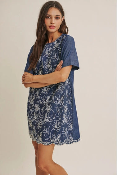 A woman wearing a short-sleeve blue dress with a white floral embroidered pattern, set against a plain beige studio background.