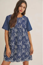 A woman wearing a short-sleeve blue dress with a white floral embroidered pattern, set against a plain beige studio background.