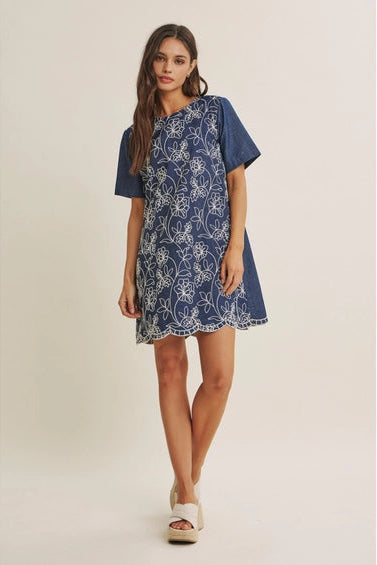 A woman wearing a short-sleeve blue dress with a white floral embroidered pattern, set against a plain beige studio background.