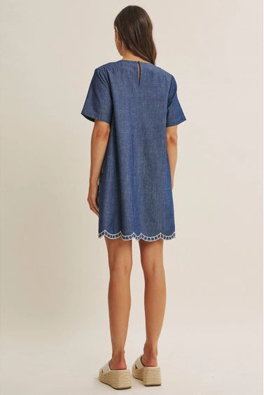 Back view of a woman wearing a short-sleeve blue dress with a white floral embroidered pattern, set against a plain beige studio background.