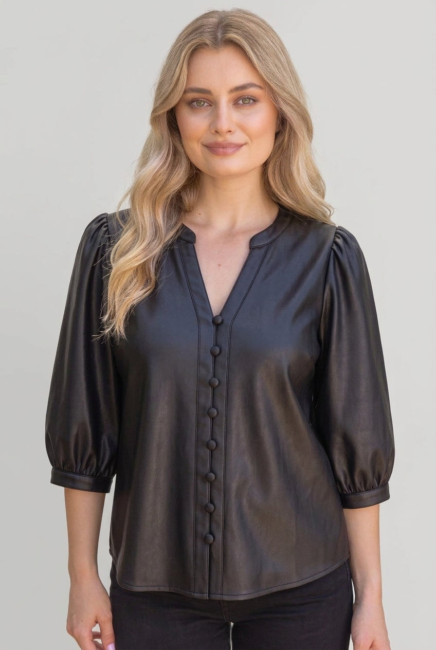 A woman wearing a black faux-leather button-up blouse with puff sleeves, standing outdoors in front of a wooden fence and green foliage.