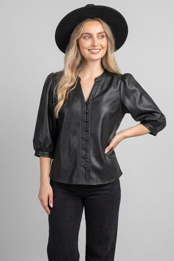 A woman wearing a black faux-leather button-up blouse with puff sleeves, standing outdoors in front of a wooden fence and green foliage.
