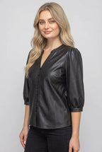 A woman wearing a black faux-leather button-up blouse with puff sleeves, standing outdoors in front of a wooden fence and green foliage.