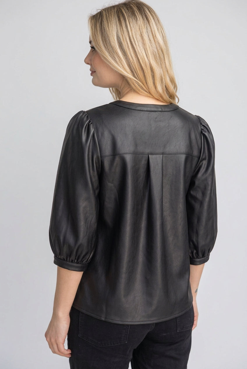 A back view of a woman wearing a black faux-leather button-up blouse with puff sleeves, standing outdoors in front of a wooden fence and green foliage.