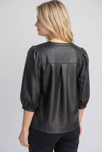 A back view of a woman wearing a black faux-leather button-up blouse with puff sleeves, standing outdoors in front of a wooden fence and green foliage.