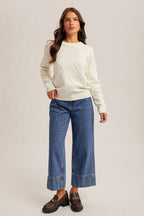 Woman wearing diamond design pearl embellishment cream sweater with jeans on gray background