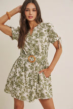 A woman wearing a short-sleeve dress with an all-over green tropical leaf print, featuring a matching belt with a round wooden buckle, set against a plain beige background.