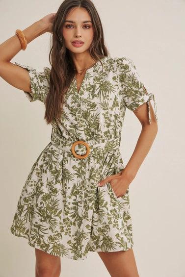 A woman wearing a short-sleeve dress with an all-over green tropical leaf print, featuring a matching belt with a round wooden buckle, set against a plain beige background.