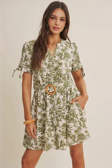 A woman wearing a short-sleeve dress with an all-over green tropical leaf print, featuring a matching belt with a round wooden buckle, set against a plain beige background.
