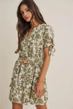 A woman wearing a short-sleeve dress with an all-over green tropical leaf print, featuring a matching belt with a round wooden buckle, set against a plain beige background.