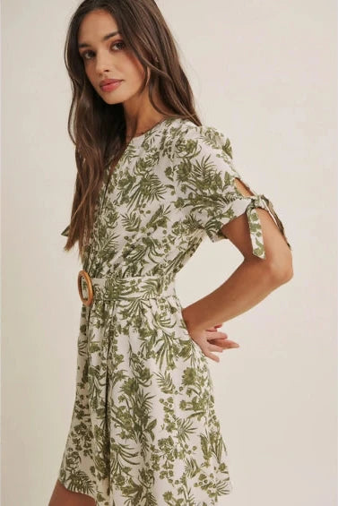 A woman wearing a short-sleeve dress with an all-over green tropical leaf print, featuring a matching belt with a round wooden buckle, set against a plain beige background.