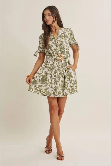 A woman wearing a short-sleeve dress with an all-over green tropical leaf print, featuring a matching belt with a round wooden buckle, set against a plain beige background.