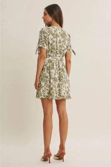 Back view of a woman wearing a short-sleeve dress with an all-over green tropical leaf print set against a plain beige background.