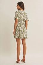 Back view of a woman wearing a short-sleeve dress with an all-over green tropical leaf print set against a plain beige background.