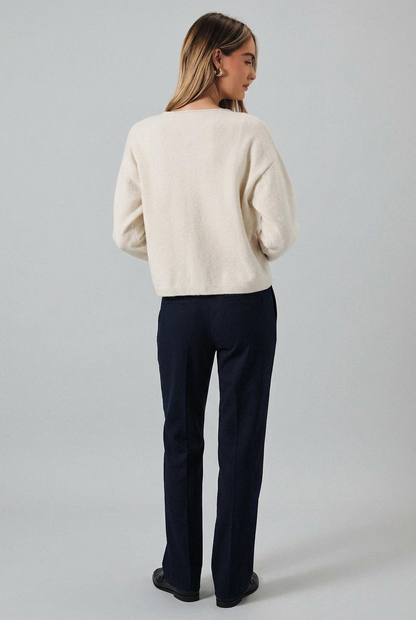 A woman shown from the back wearing a cream-colored knit sweater and dark denim shorts against a light gray studio background.