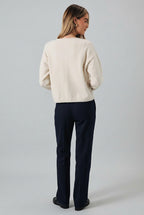 A woman shown from the back wearing a cream-colored knit sweater and dark denim shorts against a light gray studio background.